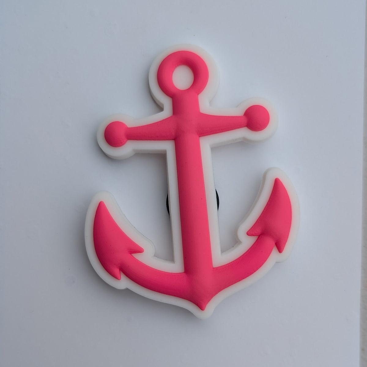 A pink anchor-shaped item with a white outline, photographed on a plain white square card against a light gray surface.
