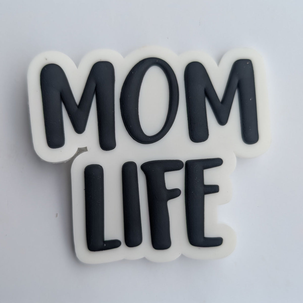 A small white rectangular card holding a raised black-and-white sticker or patch that reads “MOM LIFE” in bold rounded letters, photographed against a light neutral background.