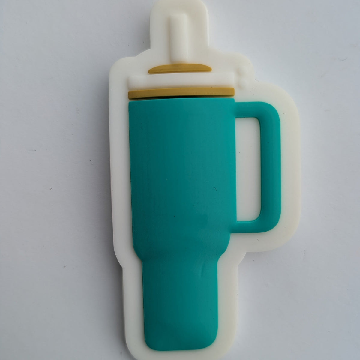 A silicone kitchen utensil shaped like a bottle, featuring a teal green body, a white outer border, and a small gold accent ring near the top, resting on a white surface.