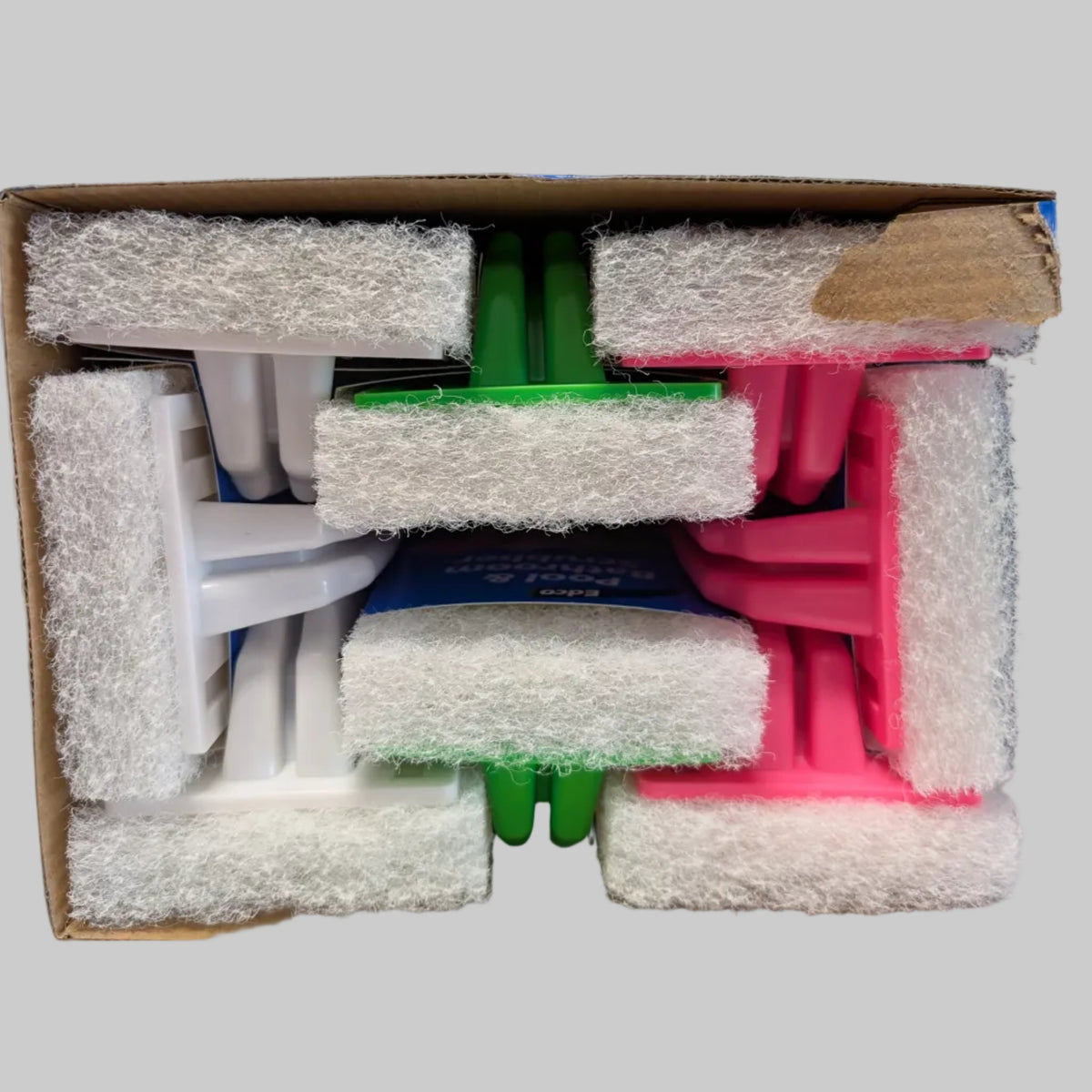 A cardboard box containing several folded white and colored plastic cleaning sponges or scrubbers wrapped in white foam padding, photographed from above on a white surface with a blurred background.