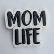A small white rectangular card holding a raised black-and-white sticker or patch that reads “MOM LIFE” in bold rounded letters, photographed against a light neutral background.